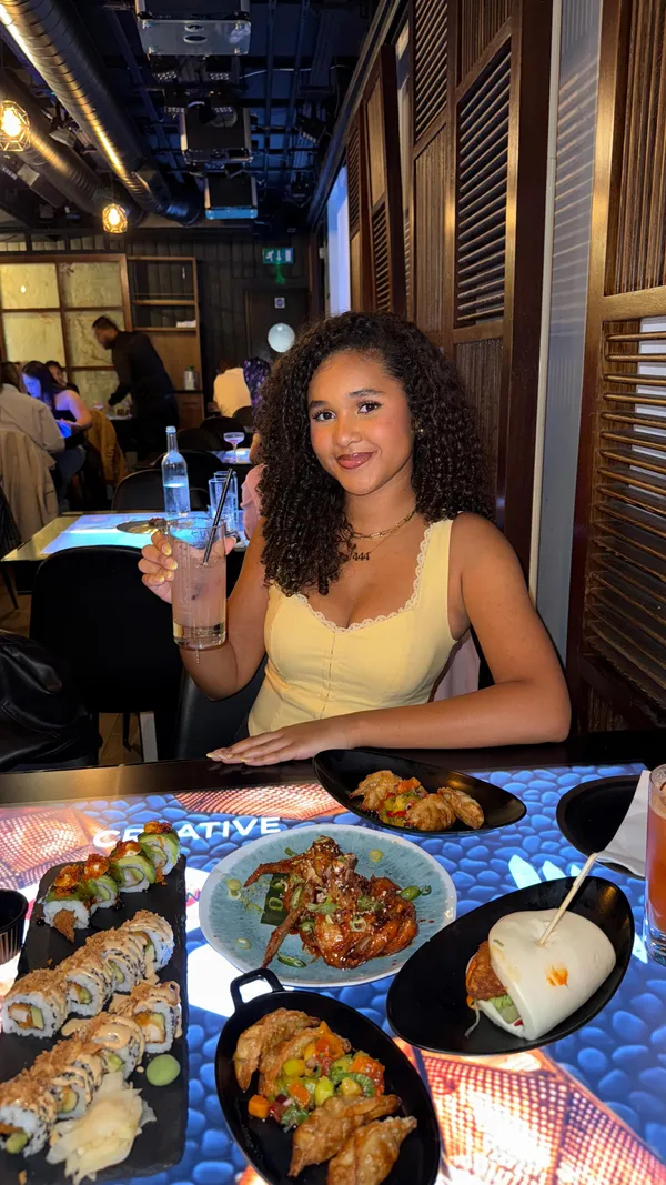 The video features a person sitting at a table in a restaurant with an interactive tabletop. Several dishes are displayed on the table, including sushi rolls, fried chicken or shrimp, dumplings, and a bao bun. The person interacts with the food, points at a dish, and takes a photo of the food with their phone. The video includes a text overlay.