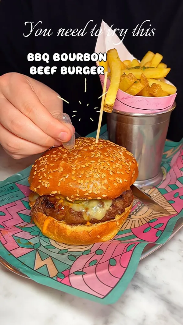 The video showcases a meal at Bills restaurant in Milton Keynes. It features a BBQ Bourbon Beef Burger with fries, a Chicken Shawarma dish with flatbread and salad, and drinks (beer and a pink cocktail, likely the Strawberry Margarita mentioned in the caption). The video shows the food presentation, people enjoying the meal, and glimpses of the restaurant's exterior and interior, including the bar and dining area. Text overlays highlight the burger and the occasion ("Friday nights").