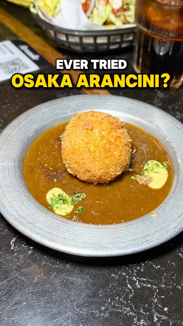 The video showcases various food items available at Arcade Food Hall in London. It starts with a close-up of an Osaka Arancini from Katsu Kamon, showing a cheese pull when cut with a fork, described as filled with melted cheese and swimming in Katsu sauce. It then shows shots of the food hall interior, a large shawarma spit, a burger, dumplings in sauce, fried chicken being dipped in sauce, and another dish of fried chicken with crispy toppings. Text overlays identify the arancini, its filling and sauce, mention Katsu Kamon as a vendor, highlight the food hall as a group dining destination, and provide the location (Arcade Tottenham Court Road).