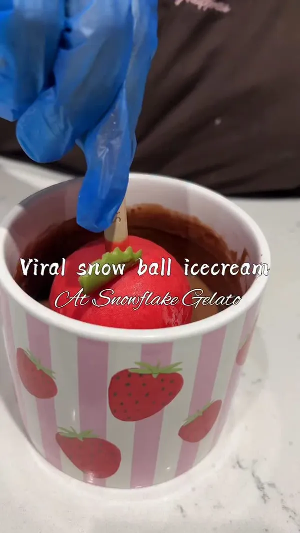 The video promotes Snowflake Gelato, focusing on their fruit-shaped gelato pops. It begins with a close-up of a strawberry-shaped gelato pop being dipped in chocolate. The video then shows the storefront, a display case of various gelato options, and three specific products on plates: a strawberry 'Snow berry', a mango-shaped pop, and a pistachio bar. The highlight is a satisfying shot of the strawberry gelato being cracked open with a spoon to reveal the pink ice cream inside.