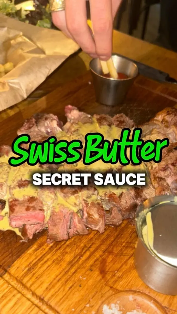 A close-up shot shows a sliced tomahawk steak on a wooden board. A hand pours a green-yellow sauce from a small metal jug over the steak. Salt is visible on the board.