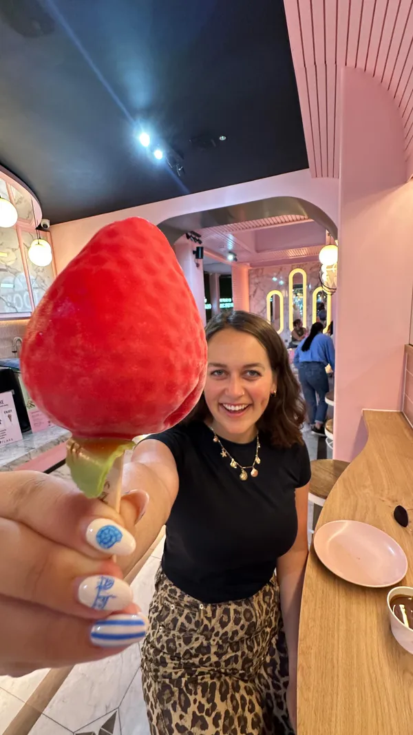 A woman is featured in a pink-themed dessert shop, holding a strawberry-shaped gelato pop with a chocolate-dipped top. The video captures her smiling at the dessert before taking a bite and showing a pleasantly surprised reaction. The content is presented as a humorous meme with a text overlay.